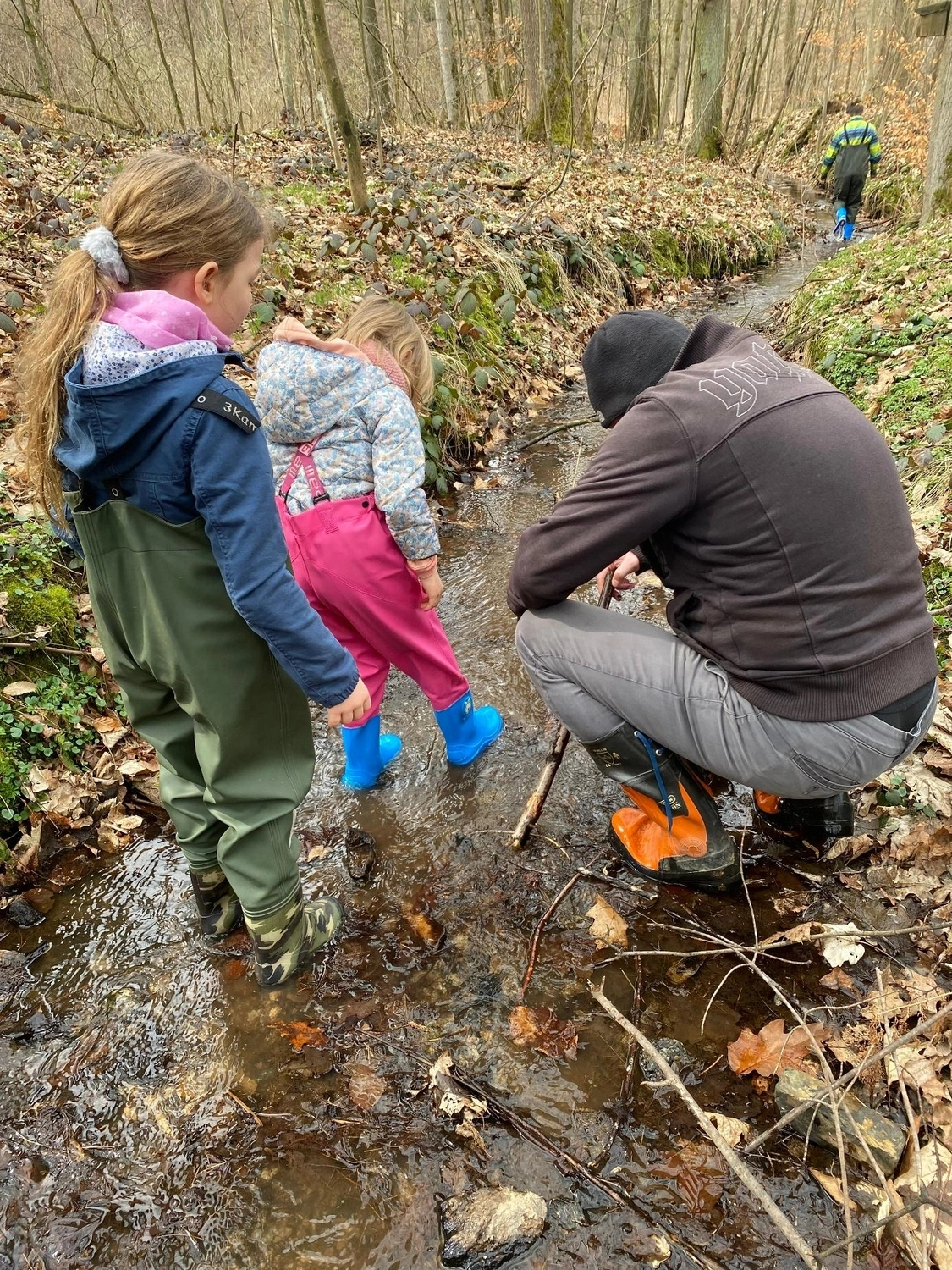 Netzwerk Colditz e.V. - Beziehung. Gestaltung. Entwicklung - Eine Person und zwei Kinder erkunden einen flachen, steinigen Bach, der von Bäumen umgeben ist. Sie tragen Outdoor-Kleidung und wasserdichte Stiefel und beobachten das Wasser in einer natürlichen, bewaldeten Umgebung.